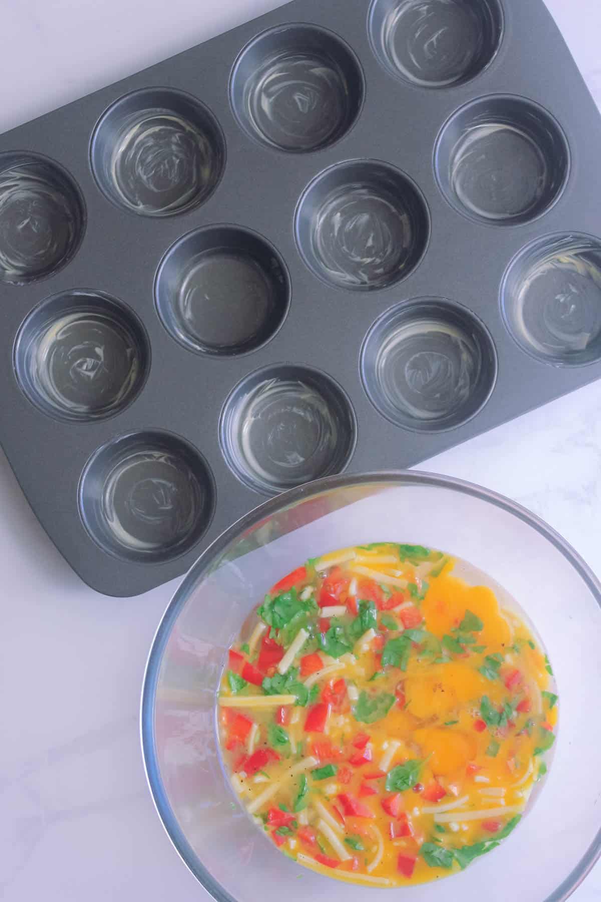Mixed egg muffin ingredients in a glass bowl with a greased muffin tray.