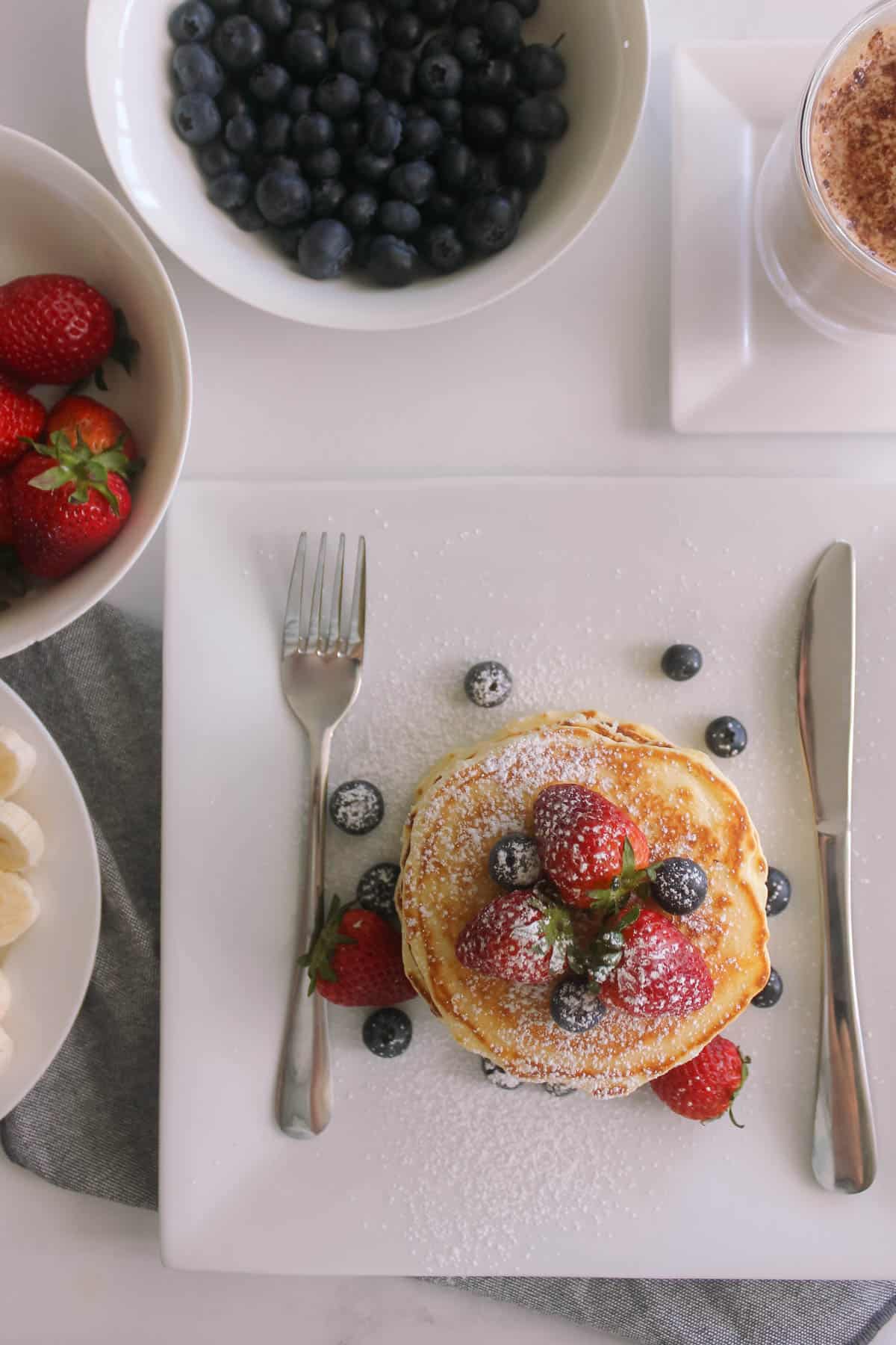 Pancakes on a plate with knife and fork, with strawberries, blueberries and coffee in background.
