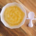 Sweet apple puree in a white bowl, displayed on a wooden chopping board with a little white spoon
