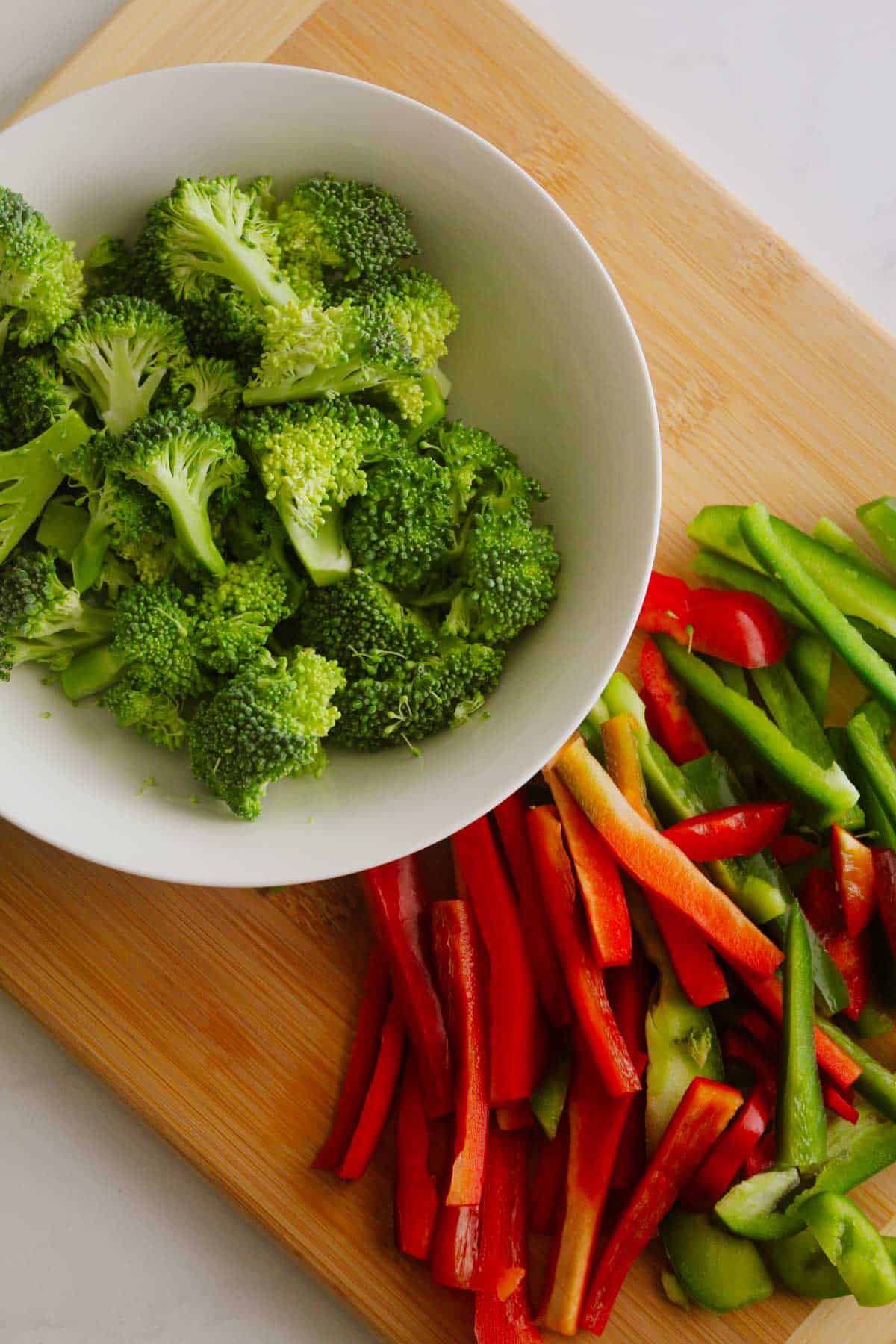 Broccoli florets in a white bowl placed on a wooden chopping board with sliced red and green capsicum