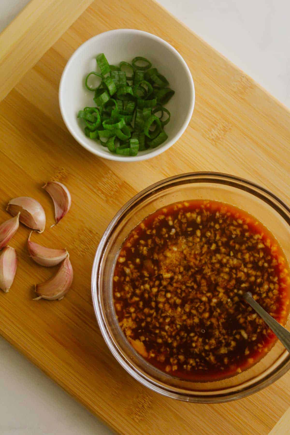 Stir-fry sauce in a glass bowl on a wooden chopping board with spring onions sliced in a white bowl and garlic cloves displayed