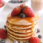 Stacked pancakes with strawberries, blueberries and maple syrup being drizzled over top.